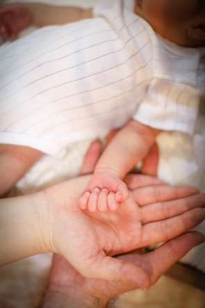 A tender moment is captured as an adult hand gently cradles the small hand of a baby, showcasing the contrast between their sizes. The baby is wearing a white outfit with thin black stripes, and the setting exudes warmth and intimacy.
