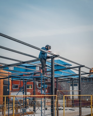 A construction worker wearing a protective helmet stands on a metal scaffold while welding steel beams. Sparks fly as the welding process creates an electric arc. The background includes a blue-roofed building and brick walls.