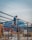 A construction worker wearing a protective helmet stands on a metal scaffold while welding steel beams. Sparks fly as the welding process creates an electric arc. The background includes a blue-roofed building and brick walls.