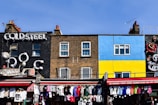 A vibrant street scene features a variety of storefronts with colorful facades. A black building with the sign 'Cold Steel' has a bold and industrial style, while an adjacent building displays a mural with bright blue and yellow sections. Below, a market stall offers an array of clothing hanging in a lively, bustling atmosphere.