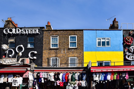 A vibrant street scene features a variety of storefronts with colorful facades. A black building with the sign 'Cold Steel' has a bold and industrial style, while an adjacent building displays a mural with bright blue and yellow sections. Below, a market stall offers an array of clothing hanging in a lively, bustling atmosphere.