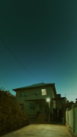 A two-story house is depicted under an evening or nighttime sky. The exterior of the house is illuminated by a single outdoor light near the entrance. There is a pathway leading to the house, flanked by dense foliage on the left side. A few windows are visible, one of which is lit. The building has a covered porch supported by columns. Electrical wires stretch across the scene.