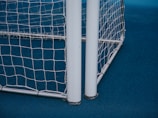 A close-up view of a soccer or football goalpost with white netting. The frame is metallic, and the ground is covered with a blue surface.