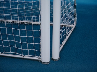 A close-up view of a soccer or football goalpost with white netting. The frame is metallic, and the ground is covered with a blue surface.