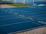 A sports track with multiple blue lanes, each marked with white numbers from 2 to 8, is visible. Adjacent to the track is a paved pathway and a green grassy area. In the background, a white soccer goal post can be seen on the field.
