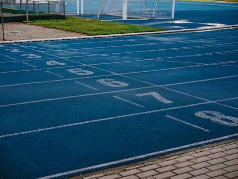 A sports track with multiple blue lanes, each marked with white numbers from 2 to 8, is visible. Adjacent to the track is a paved pathway and a green grassy area. In the background, a white soccer goal post can be seen on the field.
