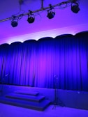 A stage is set with closed blue curtains, illuminated by overhead stage lights. Microphone stands are positioned in front of the curtains, and stairs lead up to the stage.