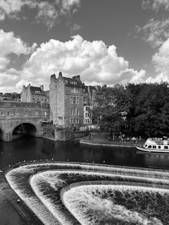 A scenic view of a historical area featuring an arched stone bridge, multi-story buildings with classic architecture, and a cascading river over a series of weirs. A tour boat is docked near the riverbank, where trees and a few people are visible. The sky is filled with large, fluffy clouds.