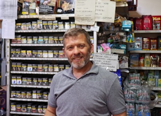 A man with short hair and a beard is standing in a store filled with shelves of various products. On the shelves behind him are packets of tobacco with health warning labels and other grocery items. There are also handwritten signs posted above the shelves.