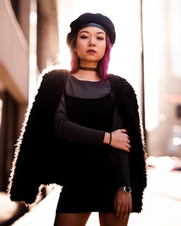 A person stands confidently in an urban setting, wearing a dark outfit and a beret. Backlit by the sun, their pink hair and choker are prominent features. The scene conveys a sense of style and individuality.