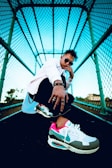 A person dressed in stylish clothing stands in a dramatic pose under a turquoise-colored metal mesh structure. The focus is on the vibrant sneakers, which feature colors like pink, teal, and white. The subject is wearing sunglasses and jewelry, projecting a confident attitude.
