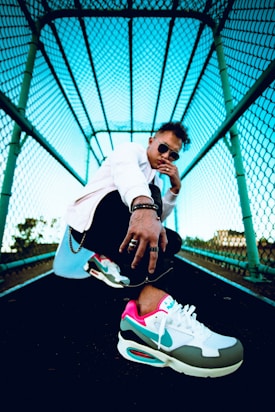 A person dressed in stylish clothing stands in a dramatic pose under a turquoise-colored metal mesh structure. The focus is on the vibrant sneakers, which feature colors like pink, teal, and white. The subject is wearing sunglasses and jewelry, projecting a confident attitude.