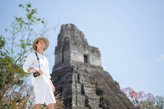 A person wearing a light-colored outfit and a hat stands in a natural setting with a digital camera hanging around their neck. Behind them is an ancient stone pyramid structure, with lush greenery and some flowering trees visible in the background. The sky is clear and bright.