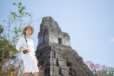A person wearing a light-colored outfit and a hat stands in a natural setting with a digital camera hanging around their neck. Behind them is an ancient stone pyramid structure, with lush greenery and some flowering trees visible in the background. The sky is clear and bright.