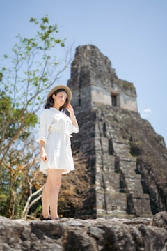 A woman in a white dress and wide-brimmed hat stands confidently near an ancient stone pyramid structure, under a clear blue sky. Sparse greenery and small trees are visible around the ruins.