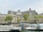 A grand, multi-story historical building with intricate architectural details, featuring large windows and ornate rooftop elements. In front, a river or canal with tourist boats docked, identified by the word 'Batobus'. Several flags, including the French and European Union flags, are visible near the water. Trees with green foliage line the embankment.