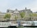 A grand, multi-story historical building with intricate architectural details, featuring large windows and ornate rooftop elements. In front, a river or canal with tourist boats docked, identified by the word 'Batobus'. Several flags, including the French and European Union flags, are visible near the water. Trees with green foliage line the embankment.