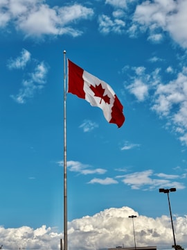 A Canadian flag with a red maple leaf in the center is flying on a flagpole against a bright blue sky scattered with fluffy white clouds. The flag is unfurled, and the scene includes the silhouettes of several streetlights below the clouds.