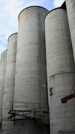Tall, cylindrical concrete silos are lined up against a partly cloudy sky. Metal walkways and fixtures are attached to the structures, contributing to an industrial appearance.
