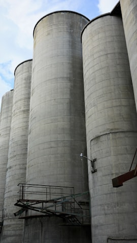 Tall, cylindrical concrete silos are lined up against a partly cloudy sky. Metal walkways and fixtures are attached to the structures, contributing to an industrial appearance.