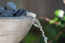 A stone basin filled with smooth black stones, with water gently flowing over the edge, surrounded by a blurred natural background.