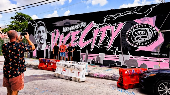 A vibrant mural on a building wall features large text saying 'Welcome to Vice City' alongside a monochromatic portrait of a person. The mural is predominantly black, pink, and gray with city skyline elements and a logo on the right side reading 'Vice City 1896 Miami.' Three people stand in front of the mural, posing for a photograph being taken by another person.