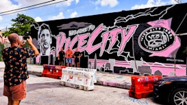 A vibrant mural on a building wall features large text saying 'Welcome to Vice City' alongside a monochromatic portrait of a person. The mural is predominantly black, pink, and gray with city skyline elements and a logo on the right side reading 'Vice City 1896 Miami.' Three people stand in front of the mural, posing for a photograph being taken by another person.