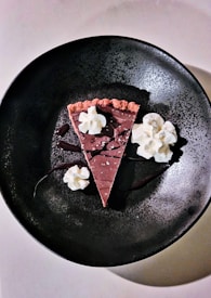 A slice of chocolate dessert on a dark, textured plate, garnished with dollops of whipped cream and drizzles of chocolate sauce. The pastry has a rich, creamy appearance, and the dessert seems elegantly presented.