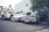Several classic cars, including a Volvo station wagon, are parked along a street. A few men stand nearby examining the vehicles. The setting appears to be urban, with buildings and a tree in the background, creating a casual and slightly nostalgic atmosphere.