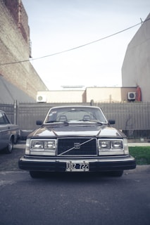 A vintage Volvo car parked on a street in an urban setting, flanked by buildings and a metal fence. The front view of the car shows its classic design, with square headlights and a distinctive grille. The license plate reads UDZ 722.