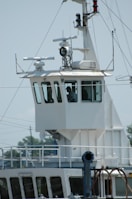 A close-up view of the bridge of a ship, featuring a control room with large windows. Various antennas and navigational equipment are mounted on the structure, and a person is visible inside the room. The ship's exterior is white, and the background shows a clear sky and blurry outlines of trees and electrical wires.
