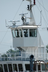 A close-up view of the bridge of a ship, featuring a control room with large windows. Various antennas and navigational equipment are mounted on the structure, and a person is visible inside the room. The ship's exterior is white, and the background shows a clear sky and blurry outlines of trees and electrical wires.