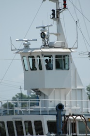 A close-up view of the bridge of a ship, featuring a control room with large windows. Various antennas and navigational equipment are mounted on the structure, and a person is visible inside the room. The ship's exterior is white, and the background shows a clear sky and blurry outlines of trees and electrical wires.