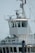 A close-up view of the bridge of a ship, featuring a control room with large windows. Various antennas and navigational equipment are mounted on the structure, and a person is visible inside the room. The ship's exterior is white, and the background shows a clear sky and blurry outlines of trees and electrical wires.
