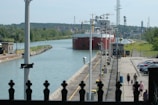 A large cargo ship is navigating through a canal lock, surrounded by green trees and industrial structures. The canal is flanked by a concrete walkway where a few people are walking near parked cars. Various utility poles and equipment are visible along the path.