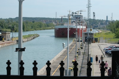 A large cargo ship is navigating through a canal lock, surrounded by green trees and industrial structures. The canal is flanked by a concrete walkway where a few people are walking near parked cars. Various utility poles and equipment are visible along the path.