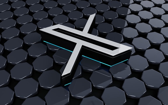 A metallic three-dimensional logo is positioned above a hexagonal tiled surface. The tiles have a glossy finish with a reflective appearance, and the logo features sharp lines and edges, with a futuristic design. Subtle neon blue lighting adds depth and contrast to the composition.