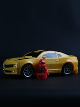 A small yellow toy car resembling a sports car is positioned on a dark surface. Next to the car, there is a red toy figurine resembling a superhero character. The car features detailed elements like wheels and side mirrors, while the figurine has detailed costume designs.