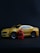 A small yellow toy car resembling a sports car is positioned on a dark surface. Next to the car, there is a red toy figurine resembling a superhero character. The car features detailed elements like wheels and side mirrors, while the figurine has detailed costume designs.