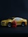 A small yellow toy car resembling a sports car is positioned on a dark surface. Next to the car, there is a red toy figurine resembling a superhero character. The car features detailed elements like wheels and side mirrors, while the figurine has detailed costume designs.