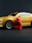 A small red figurine resembling a ninja stands in front of a toy yellow sports car with black accents. The car features detailed rims and a sleek design, set against a dark background.
