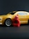 A small red figurine resembling a ninja stands in front of a toy yellow sports car with black accents. The car features detailed rims and a sleek design, set against a dark background.