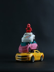 A small stack of toy cars is arranged with a red toy figure on top. The stack includes a yellow toy car at the bottom, followed by a pink and white car, topped with a red action figure. The background is a dark, solid color, emphasizing the toys.