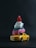 A small stack of toy cars is arranged with a red toy figure on top. The stack includes a yellow toy car at the bottom, followed by a pink and white car, topped with a red action figure. The background is a dark, solid color, emphasizing the toys.