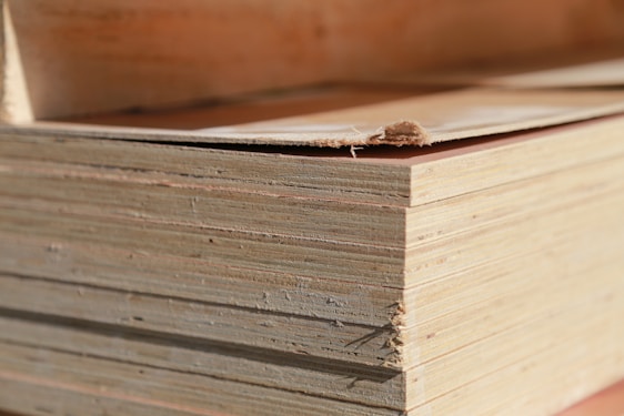 A close-up view of a stack of plywood sheets. The edges of the sheets are visible, revealing the multiple layers that make up the plywood. The top sheet has a small tear at the edge.