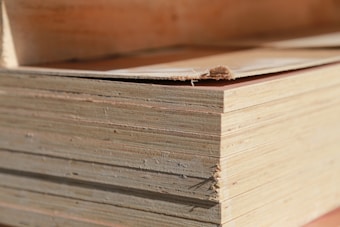 A close-up view of a stack of plywood sheets. The edges of the sheets are visible, revealing the multiple layers that make up the plywood. The top sheet has a small tear at the edge.