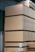 Large stacks of plywood sheets are neatly arranged in a warehouse setting. The plywood is organized in thick piles, each secured with green straps. The warehouse has a corrugated metal wall and roof, and there are labels with measurements attached to some of the stacks.