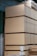 Large stacks of plywood sheets are neatly arranged in a warehouse setting. The plywood is organized in thick piles, each secured with green straps. The warehouse has a corrugated metal wall and roof, and there are labels with measurements attached to some of the stacks.