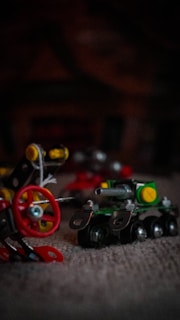 A close-up of miniature metal and plastic construction toys featuring a small toy tank with tracks and a red wheel assembly with bolts and connecting parts, set against a dark, blurred background.
