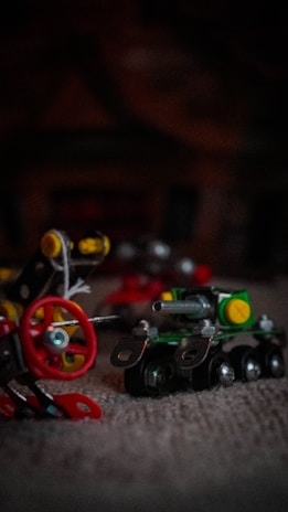 A close-up of miniature metal and plastic construction toys featuring a small toy tank with tracks and a red wheel assembly with bolts and connecting parts, set against a dark, blurred background.